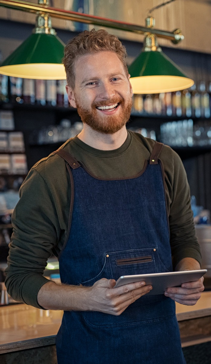 Smiling barista in a dark green shirt and blue apron holds a tablet behind a cafe counter under green pendant lights.