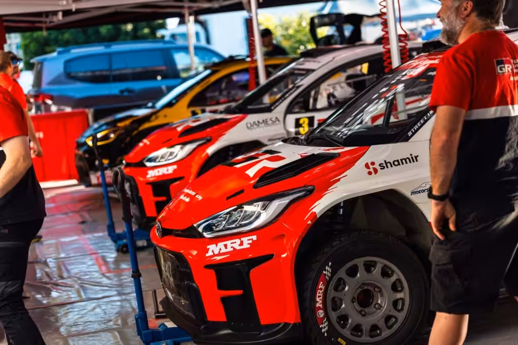 Red and white rally car in the pit area, front view with MRF branding and a mechanic nearby under a canopy.